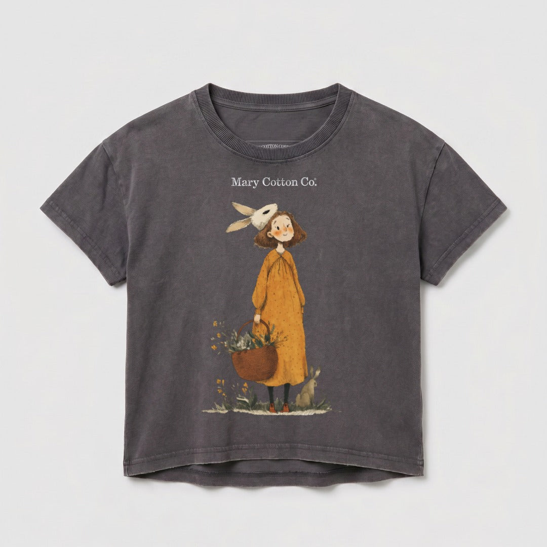 Girl with bunny ears and basket, next to a rabbit, on a dark gray t-shirt.